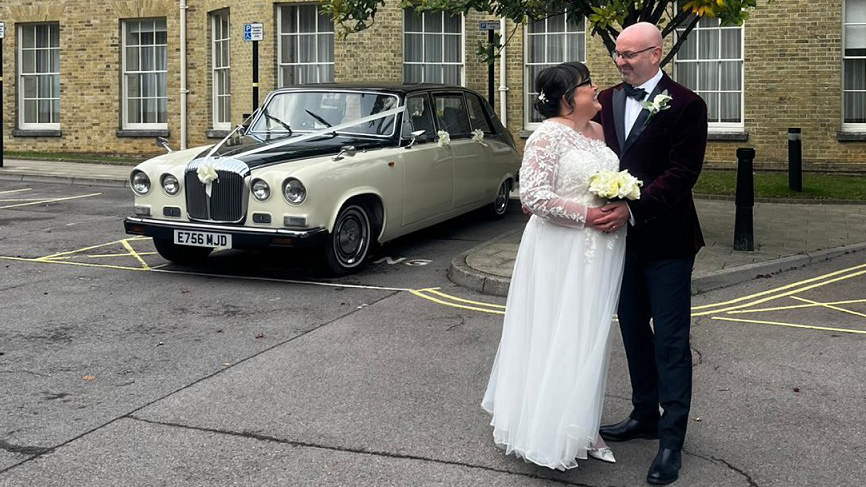 Bride in white gown and groom in dark suit standing beside a black and ivory Daimler DS420 limousine outside a stone building venue, car positioned side-on with chrome details and wedding ribbons visible on the bonnet.