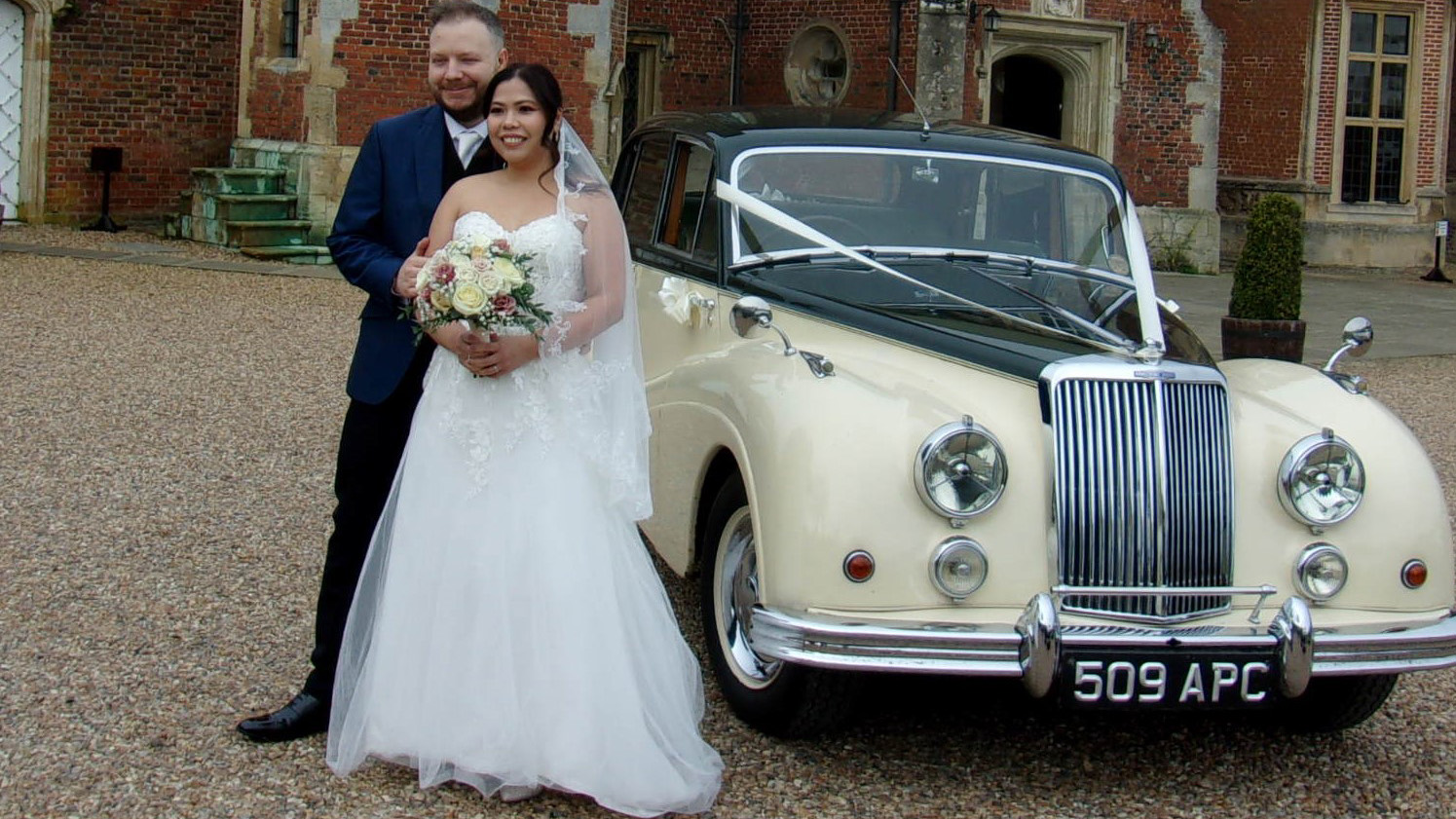 Bride and groom standing beside ivory Armstrong Siddeley with white ribbon decoration, gravel courtyard and brick building behind.