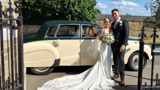 Bride and groom outside church entering ivory classic car, vehicle positioned roadside with stone building backdrop.