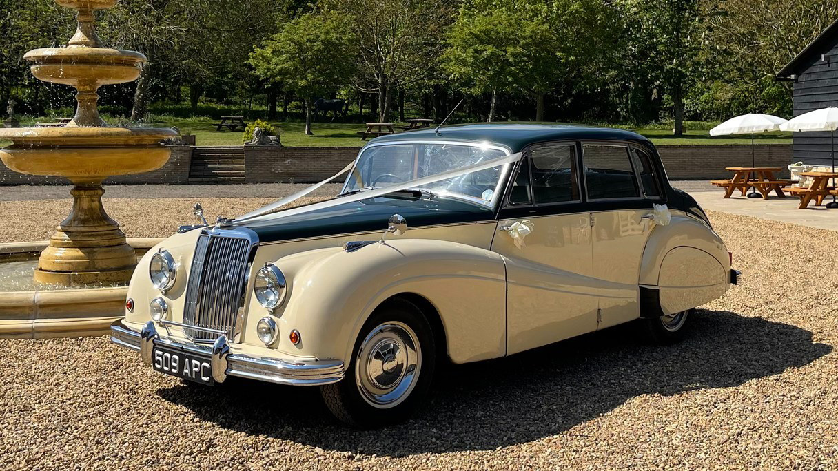 Ivory and green Armstrong Siddeley parked on gravel driveway near fountain, sunny day with formal garden surroundings.