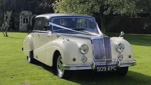 Front view of ivory Armstrong Siddeley parked on lawn with white ribbon decoration, chrome grille prominent, trees in background.