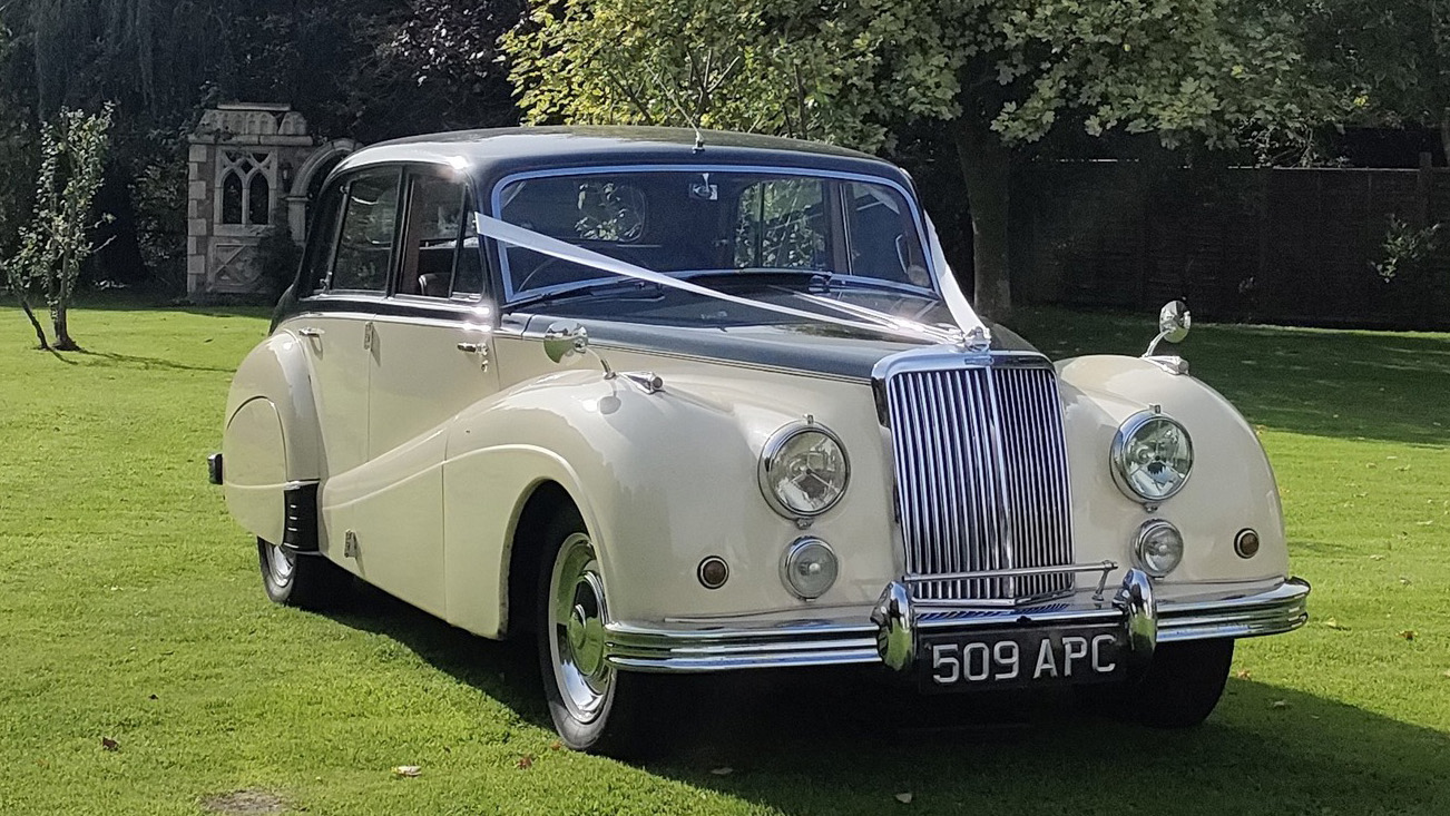 Front view of ivory Armstrong Siddeley parked on lawn with white ribbon decoration, chrome grille prominent, trees in background.