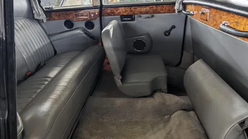 Interior rear cabin of Daimler DS420 limousine showing grey leather bench seats with one occasional fold-down seat open, positioned facing forward, wood trim panels along the sides and large window space letting in natural light.