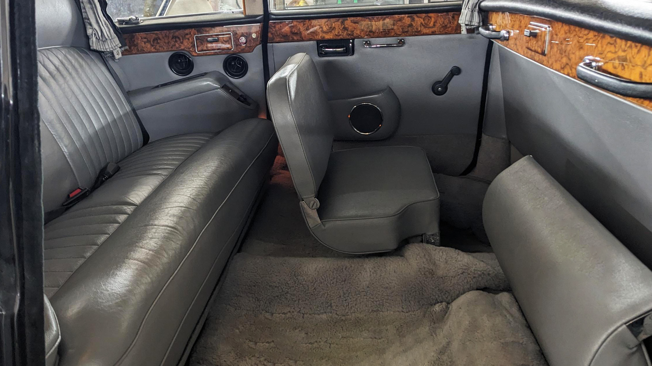 Interior rear cabin of Daimler DS420 limousine showing grey leather bench seats with one occasional fold-down seat open, positioned facing forward, wood trim panels along the sides and large window space letting in natural light.
