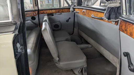 Rear cabin of Daimler DS420 with both occasional seats deployed opposite the main bench seats, creating a face-to-face seating layout, grey leather upholstery with polished wood trim and spacious legroom visible.