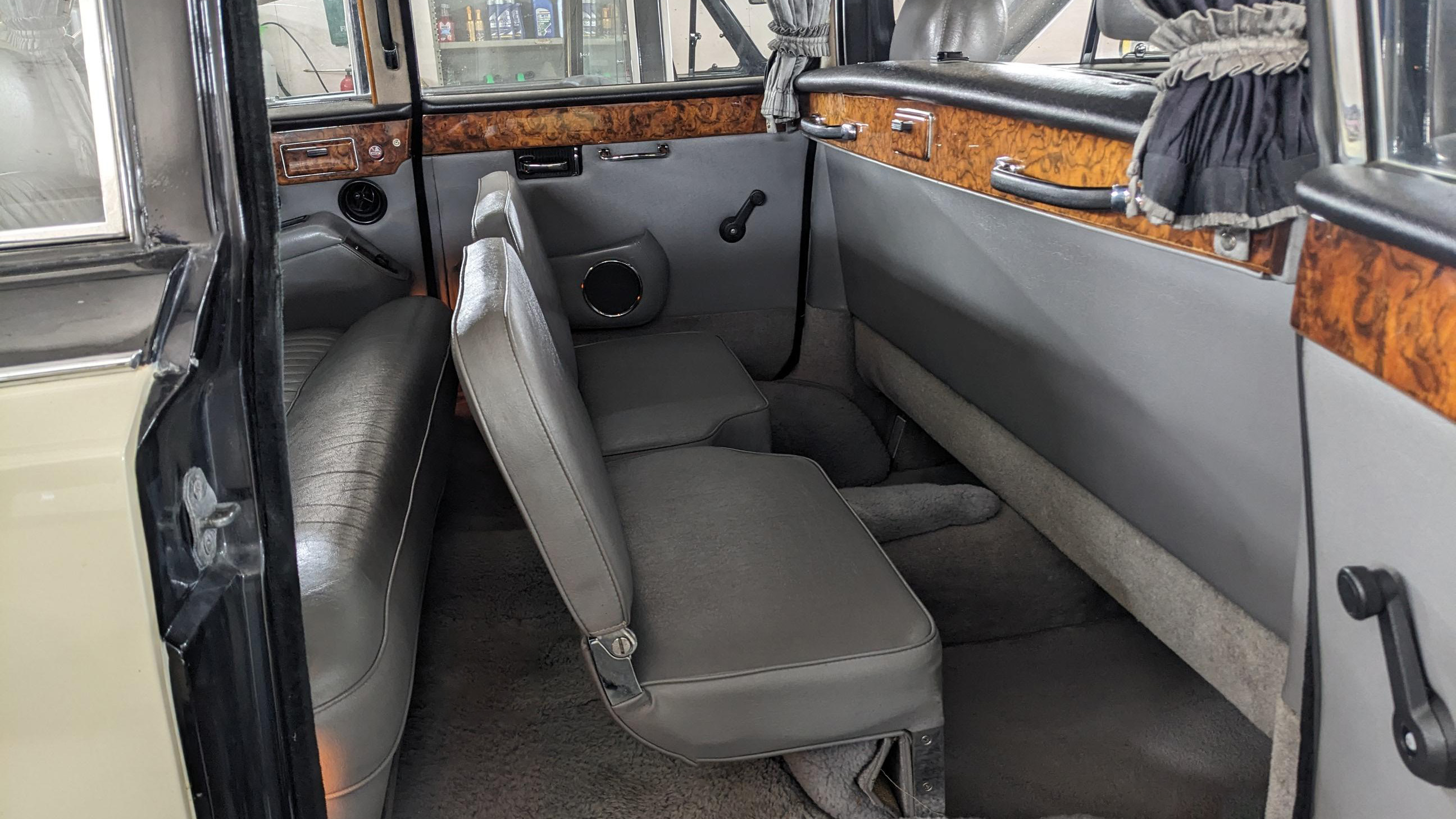 Rear cabin of Daimler DS420 with both occasional seats deployed opposite the main bench seats, creating a face-to-face seating layout, grey leather upholstery with polished wood trim and spacious legroom visible.