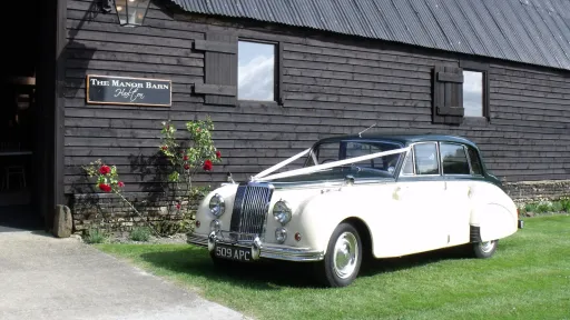 Ivory and green Armstrong Siddeley classic wedding car parked on grass beside a dark wooden barn, car is dressed with white ribbons, countryside setting.