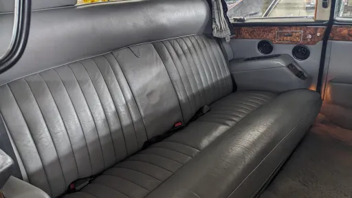 Close-up of rear bench seat inside Daimler DS420 limousine, upholstered in grey leather with vertical stitching, wood trim panels above and soft natural lighting entering through side windows.