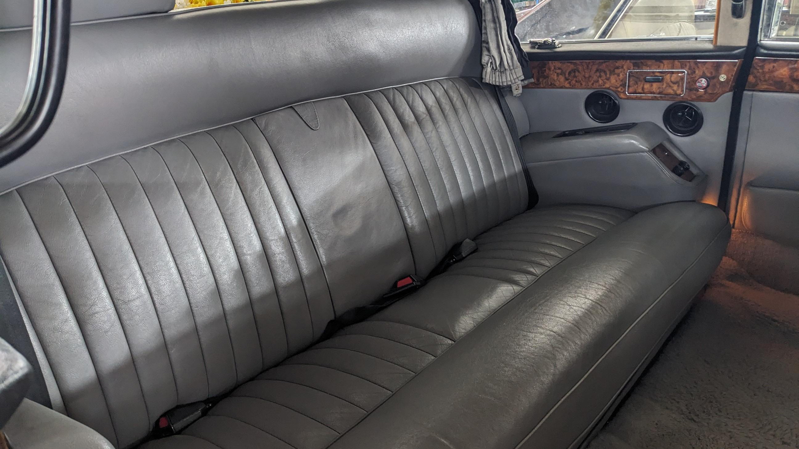 Close-up of rear bench seat inside Daimler DS420 limousine, upholstered in grey leather with vertical stitching, wood trim panels above and soft natural lighting entering through side windows.