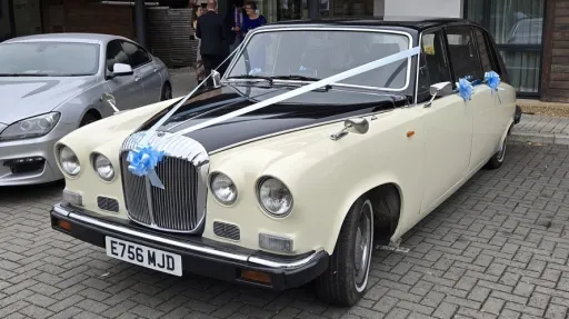 Ivory Daimler DS420 limousine parked on a paved area with white wedding ribbons across the bonnet, front angle view showing chrome grille and headlights, subtle blue ribbon accents and residential background.