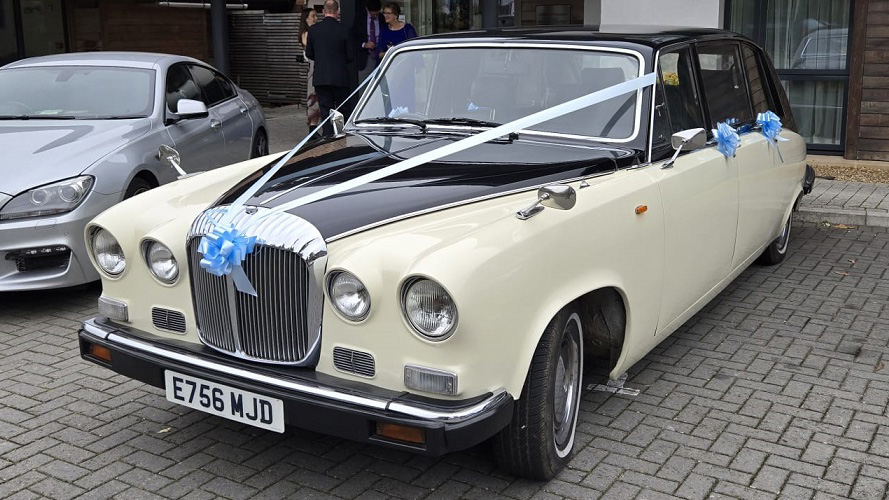 Ivory Daimler DS420 limousine parked on a paved area with white wedding ribbons across the bonnet, front angle view showing chrome grille and headlights, subtle blue ribbon accents and residential background.