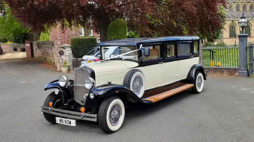 Black and ivory Bramwith limousine in 1930s vintage style, parked on road with trees and houses in background. Car is decorated with a pale Pink ribbon and Bow
