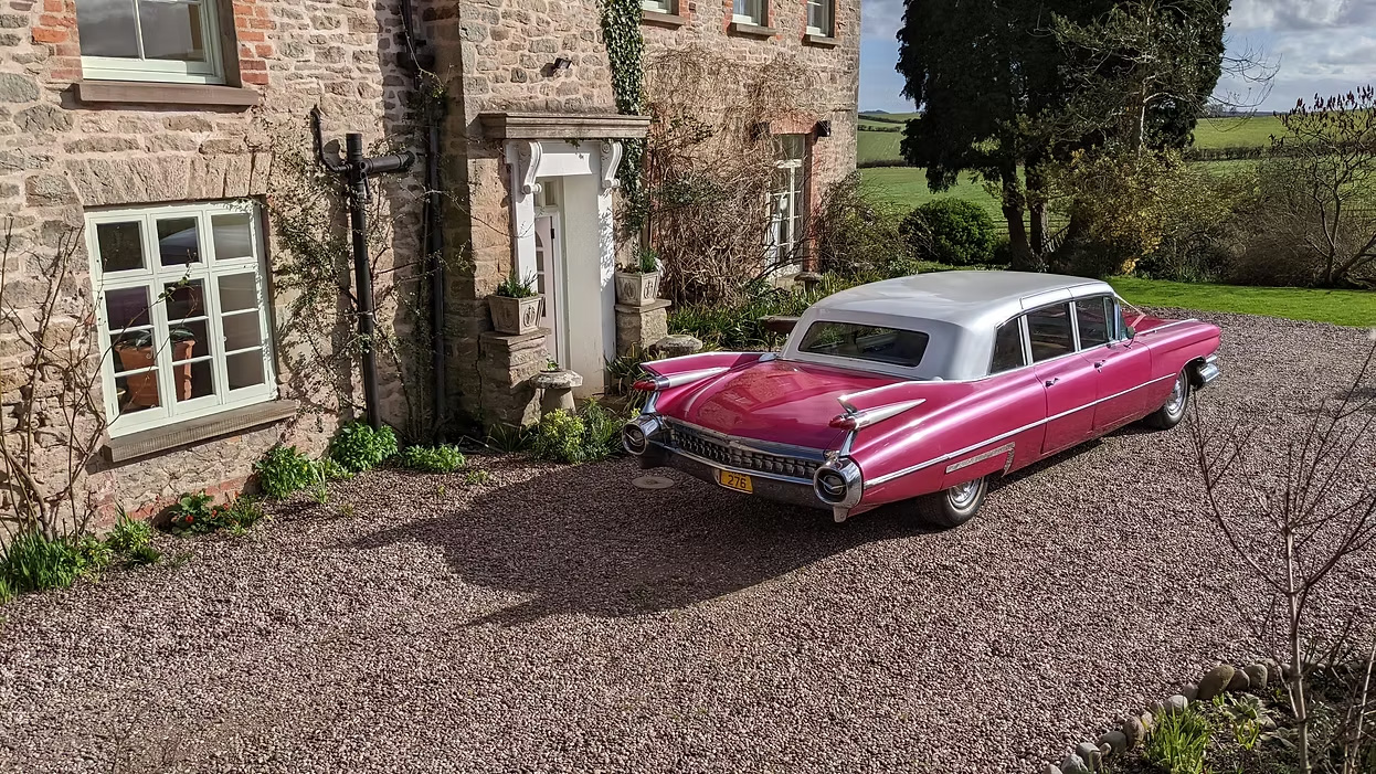 Pink Cadillac Series 75 Fleetwood limousine with white roof, viewed from rear angle on gravel driveway outside stone house, countryside background.
