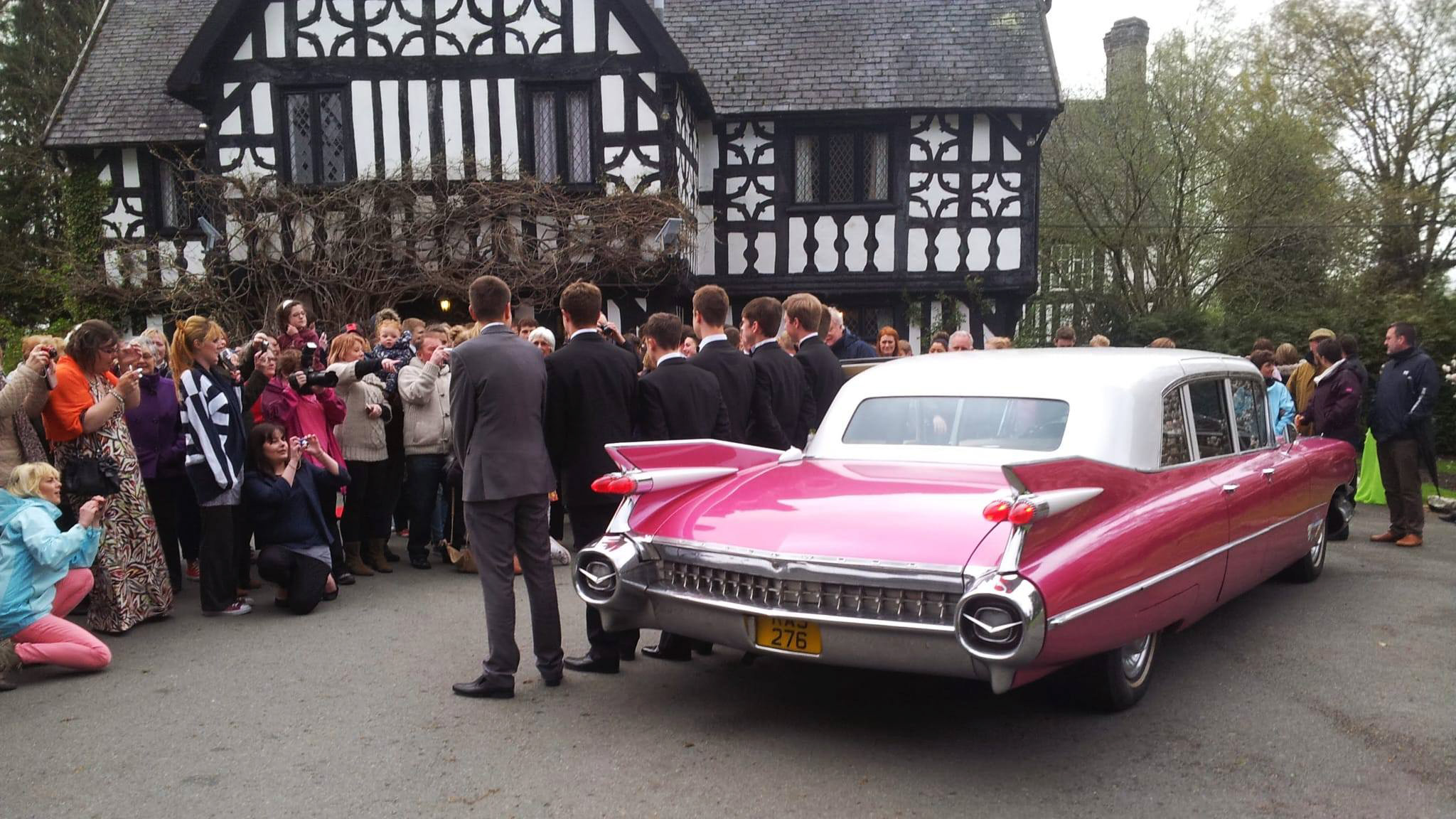 Pink Cadillac Fleetwood limousine with white roof parked outside Tudor-style venue, surrounded by wedding guests gathered near rear of car.