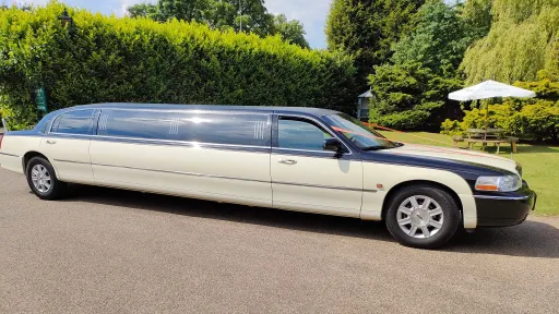 Black and ivory American Lincoln stretched limousine parked on road, full side profile with greenery and hedge background.