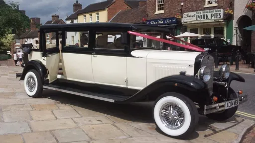 Black and ivory Bramwith Landaulette limousine with cream interior, parked on paved area with stone buildings in background dressed with burgundy red Wedding Ribbons.