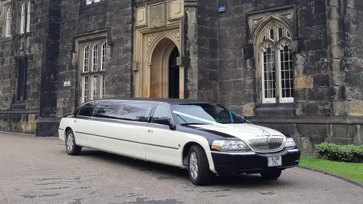 Black and ivory Lincoln stretch limousine parked outside historic stone building, front angle view with arched doorway behind.