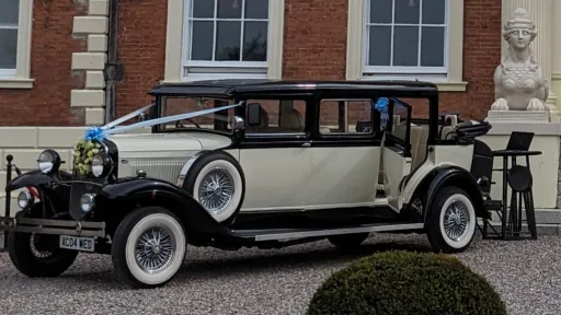 Black and ivory Bramwith Landaulette wedding car parked outside venue, side profile showing vintage styling and white wall tyres decorated with light blue ribbons and bows in front of the bonnet.