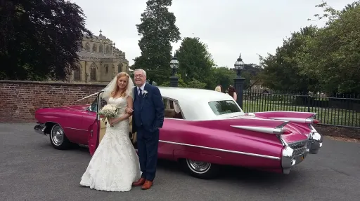 Bride and groom standing in front of pink Cadillac limousine with white roof, parked on paved area with trees and greenery behind.