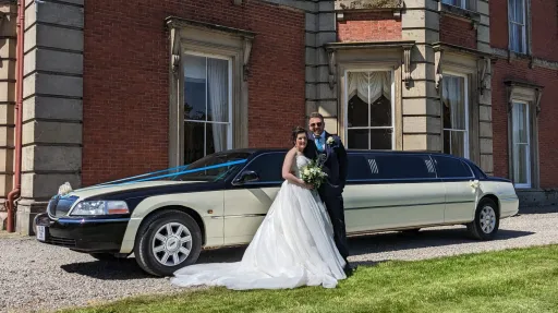 Bride and groom standing beside black and ivory Lincoln stretch limousine outside venue, full vehicle visible with building backdrop.