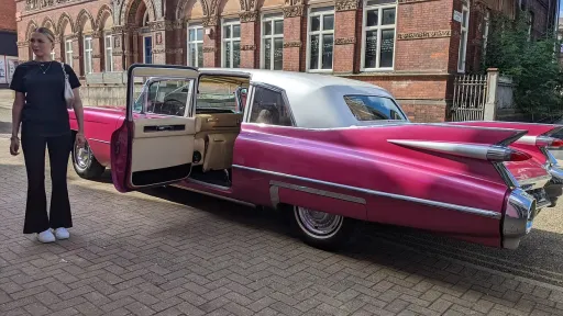 Pink Cadillac limousine with rear passenger door open, showing cream interior, parked outside brick building with chauffeur nearby.