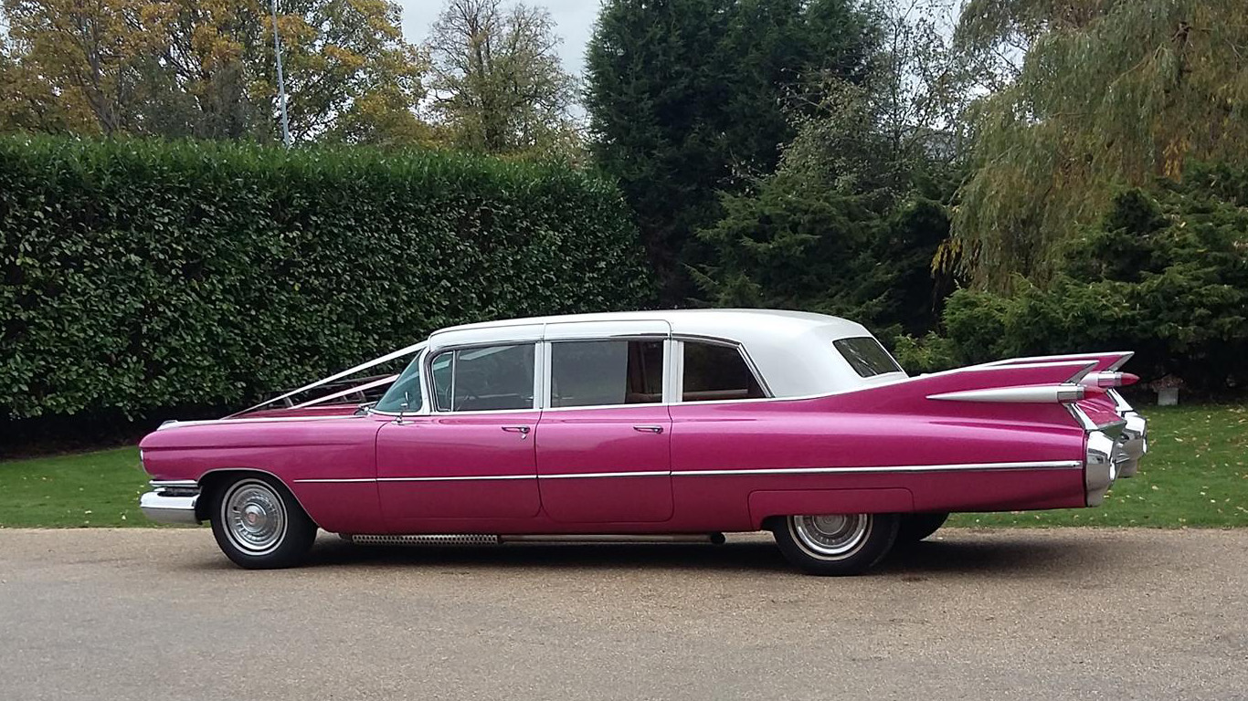 Full left side view of pink Cadillac limousine with white roof, parked on road with hedges and trees in background.