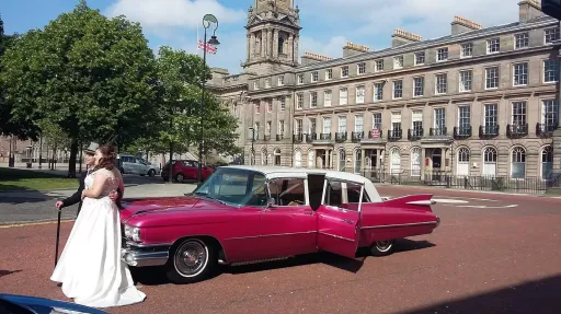 Bride standing beside pink Cadillac limousine with white roof in city square, historic buildings visible in background.