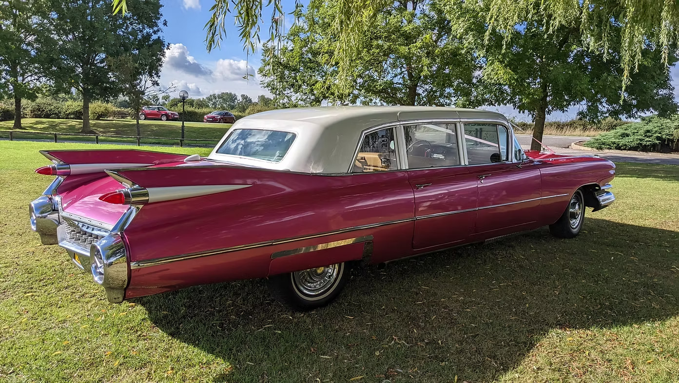 Rear right angle of pink Cadillac limousine with white roof parked on grass, chrome bumper and tail fins visible.