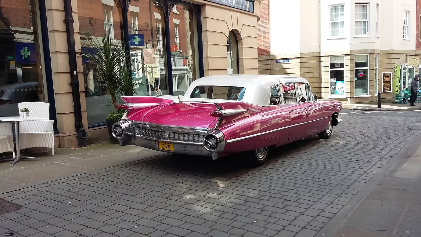 Rear view of pink Cadillac Fleetwood limousine with white roof parked on paved street outside buildings.