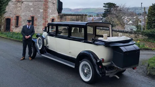 Rear view of black and ivory Bramwith Landaulette limousine with chauffeur standing in front of the vehicle, parked outside stone venue.
