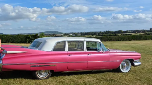 Right side profile of pink Cadillac limousine with white roof, parked on country road with open fields behind.