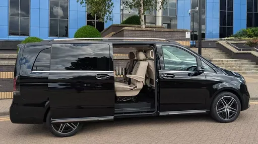 Black Mercedes V-Class limousine side profile with sliding door open, cream interior visible, parked outside glass-fronted venue.