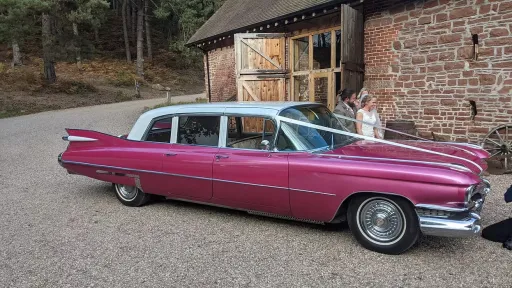 Pink Cadillac limousine parked outside rustic barn venue, right side visible with countryside surroundings.