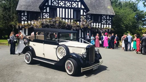 Black and ivory Cowley vintage limousine with landaulette roof, parked outside Tudor venue with wedding guests in background, white wall tyres and side mounted spare wheel visible.