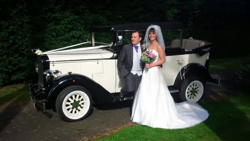 Bride and groom standing beside black and ivory Cowley landaulette limousine at dusk, convertible rear roof open, chrome detailing and white wall tyres.