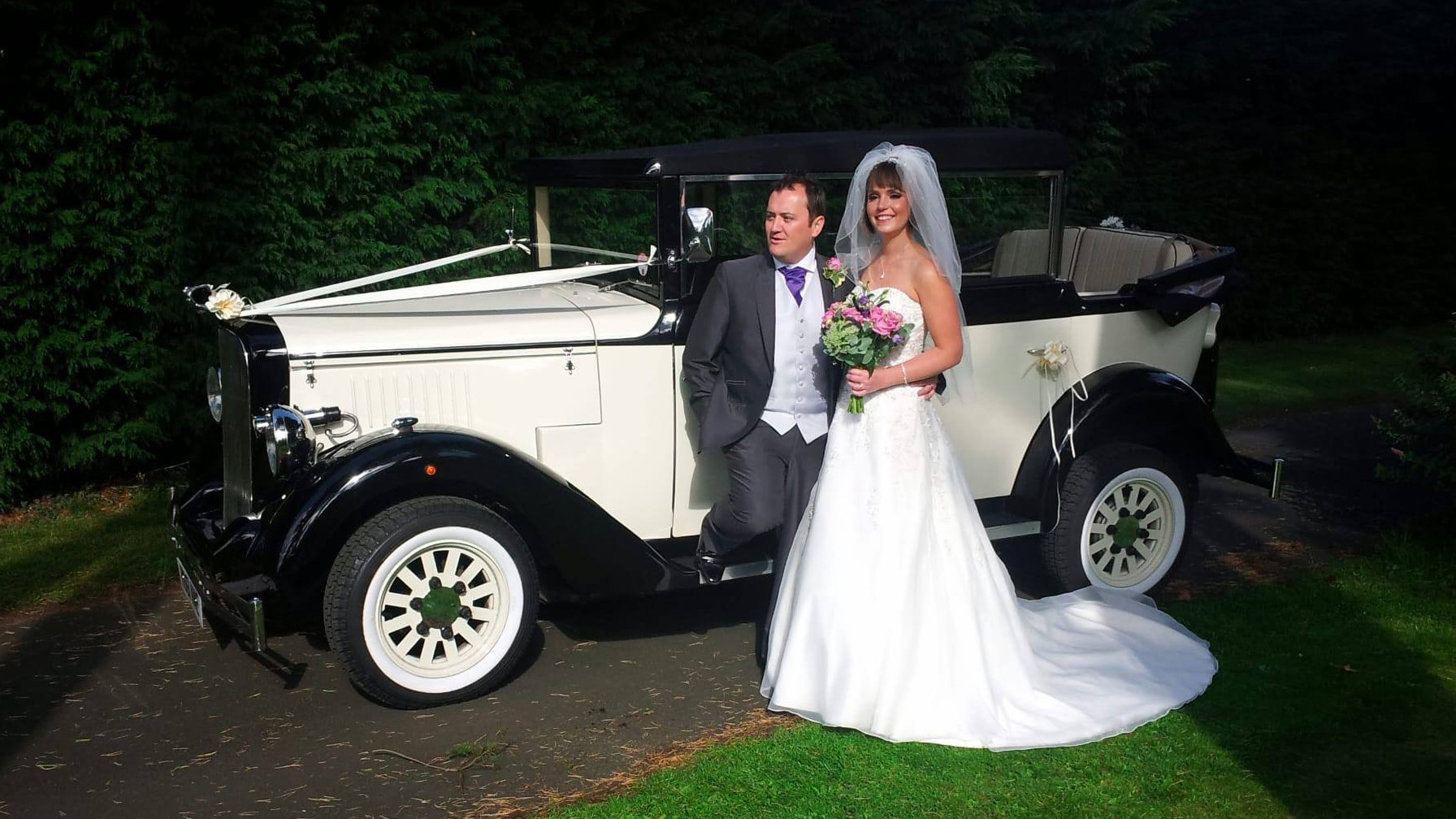 Bride and groom standing beside black and ivory Cowley landaulette limousine at dusk, convertible rear roof open, chrome detailing and white wall tyres.