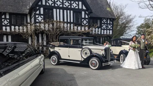 Bride and groom beside two matching black and ivory Cowley vintage limousines with landaulette roofs, parked outside Tudor-style venue.