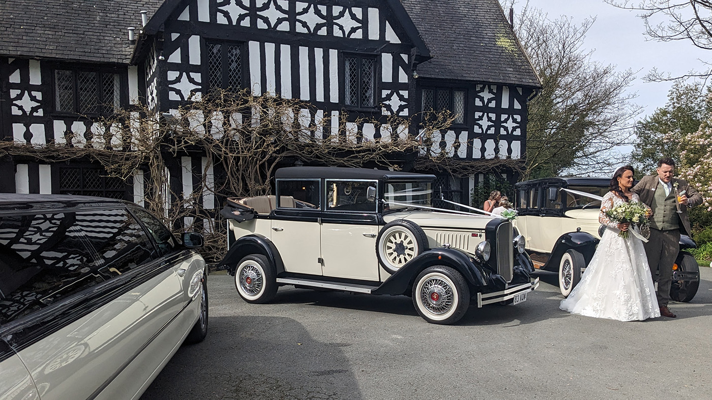 Bride and groom beside two matching black and ivory Cowley vintage limousines with landaulette roofs, parked outside Tudor-style venue.