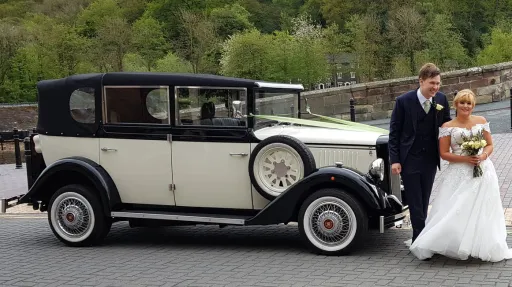 Bride and groom posing next to black and ivory Cowley landaulette wedding car, rear roof open, classic white wall tyres and side spare wheel.