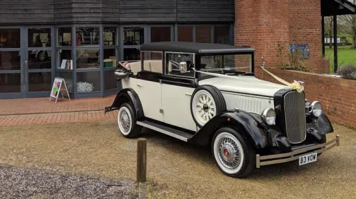 Black and ivory Cowley landaulette limousine parked outside wedding venue, rear convertible roof open, picnic trunk and side mounted spare wheel visible.