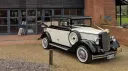 Black and ivory Cowley landaulette limousine parked outside wedding venue, rear convertible roof open, picnic trunk and side mounted spare wheel visible.