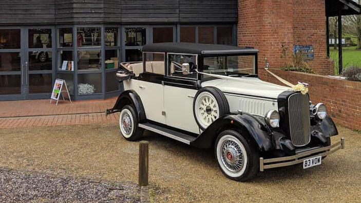 Black and ivory Cowley landaulette limousine parked outside wedding venue, rear convertible roof open, picnic trunk and side mounted spare wheel visible.