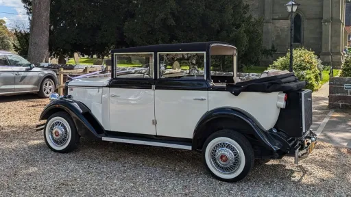 Black and ivory Cowley limousine with landaulette roof open, parked near stone building, showing long body and vintage detailing.