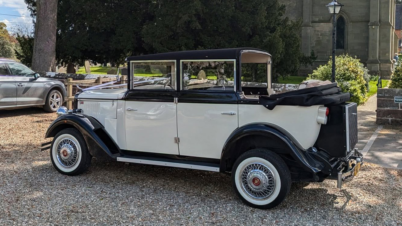 Black and ivory Cowley limousine with landaulette roof open, parked near stone building, showing long body and vintage detailing.