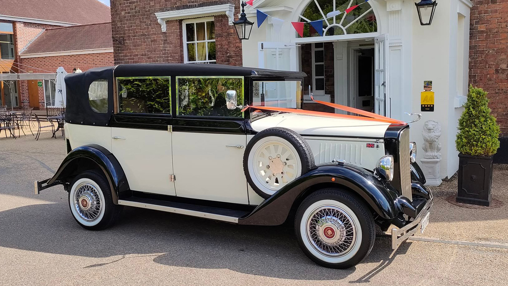 Black and ivory Cowley landaulette wedding car decorated with red ribbons on bonnet, side view outside venue with white wall tyres and sapre wheel mounted on the right wide of the vehicle