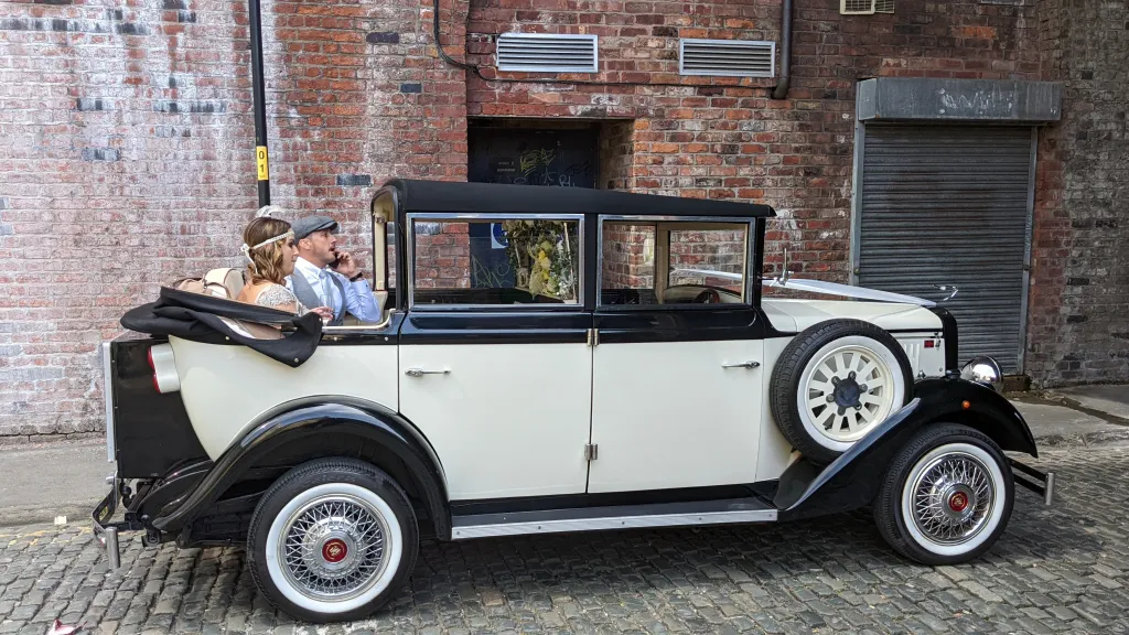 Cowley landaulette limousine with rear roof open, couple seated inside, showing picnic trunk, side spare wheel and vintage styling.