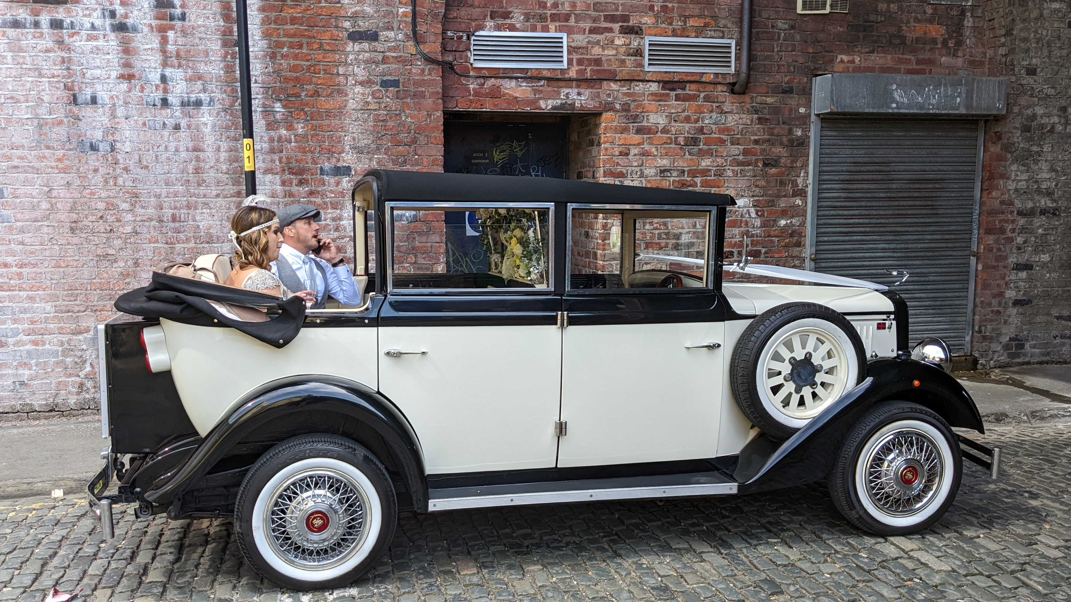 Cowley landaulette limousine with rear roof open, couple seated inside, showing picnic trunk, side spare wheel and vintage styling.