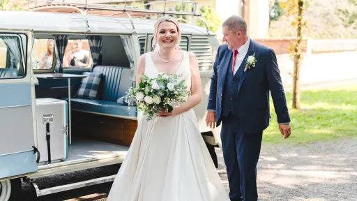 Bride and father walking beside a cream VW split screen campervan, side door open showing turquoise interior, outdoor wedding setting.