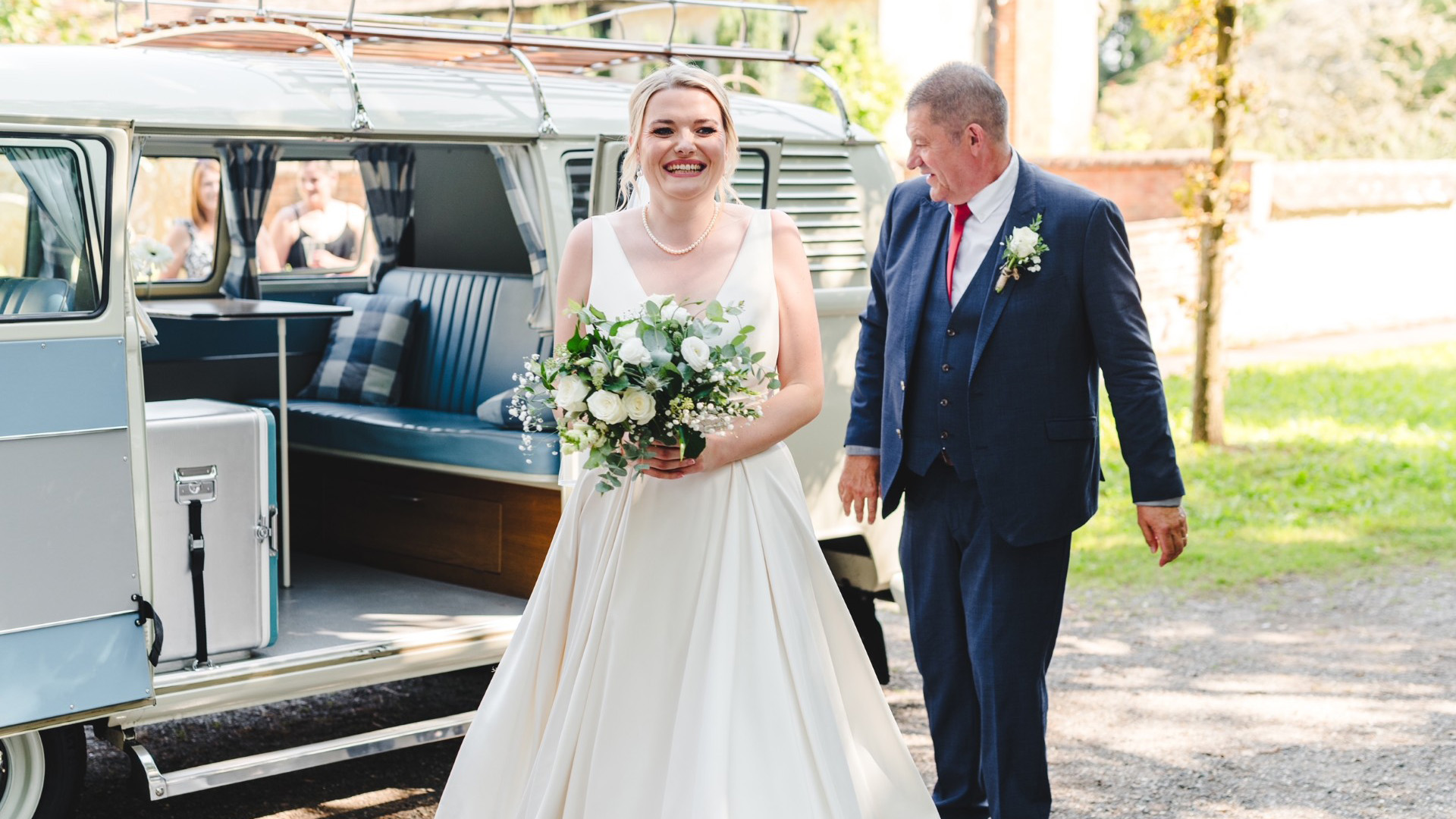 Bride and father walking beside a cream VW split screen campervan, side door open showing turquoise interior, outdoor wedding setting.