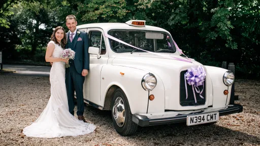 Bride and groom posing beside an Old English White classic taxi cab decorated with wedding ribbons, set on a gravel driveway.